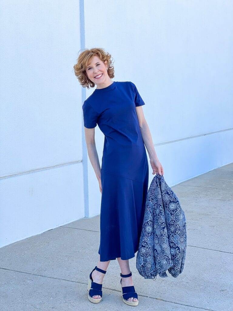 woman over 50 standing against white wall wearing Caslon® A-Line T-Shirt Cotton Maxi Dress from Nordstrom with navy wedges and holding a navy quilted patterned jacket