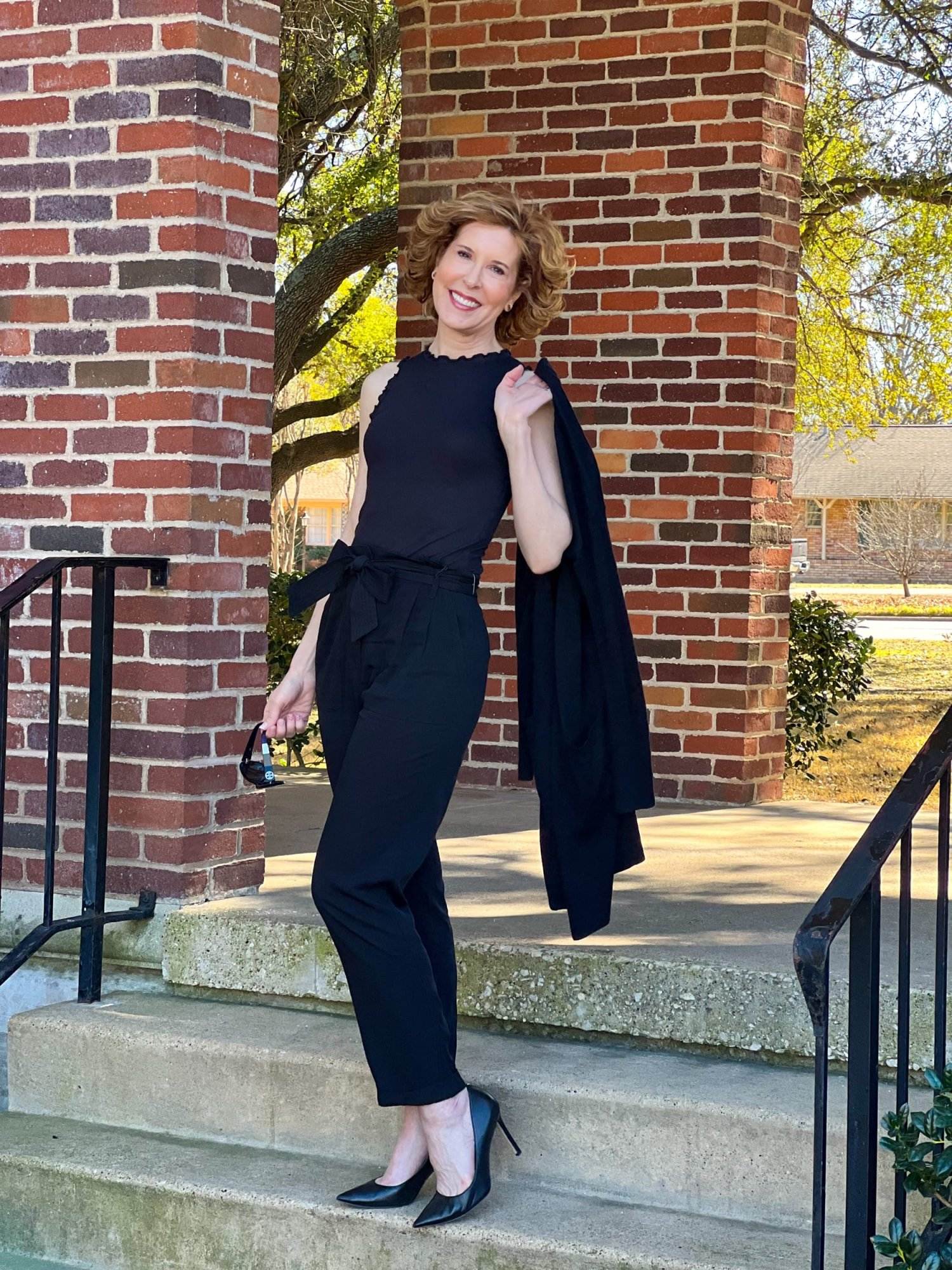 woman over 50 posing on staircase wearing black Treasure & Bond Paperbag Waist Pants, wearing Ruffle Rib Tank and an open front cardigan slung over her shoulder as she transitions her wardrobe from spring to winter