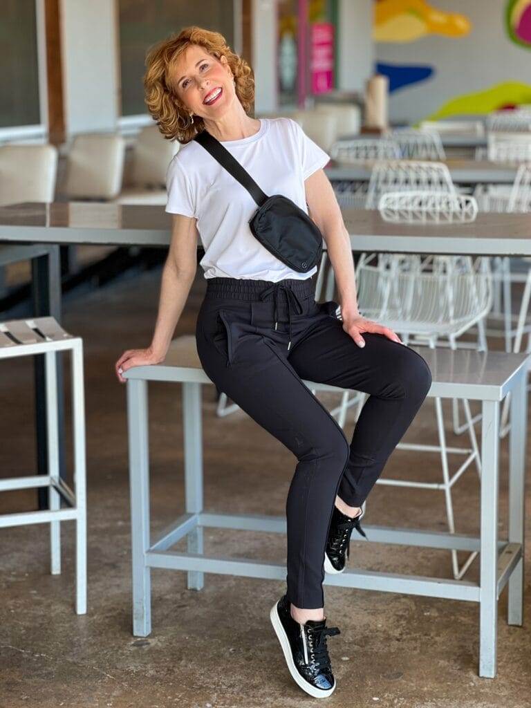 woman over 50 posing on a stool on a restaurant patio wearing black pants and a white tee with a black lululemon belt bag