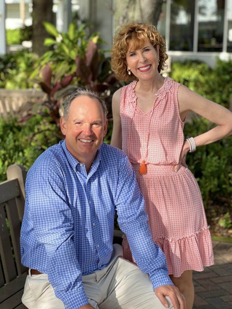 woman over 50 wearing coral dress standing alongside sitting man over 50 wearing blue shirt