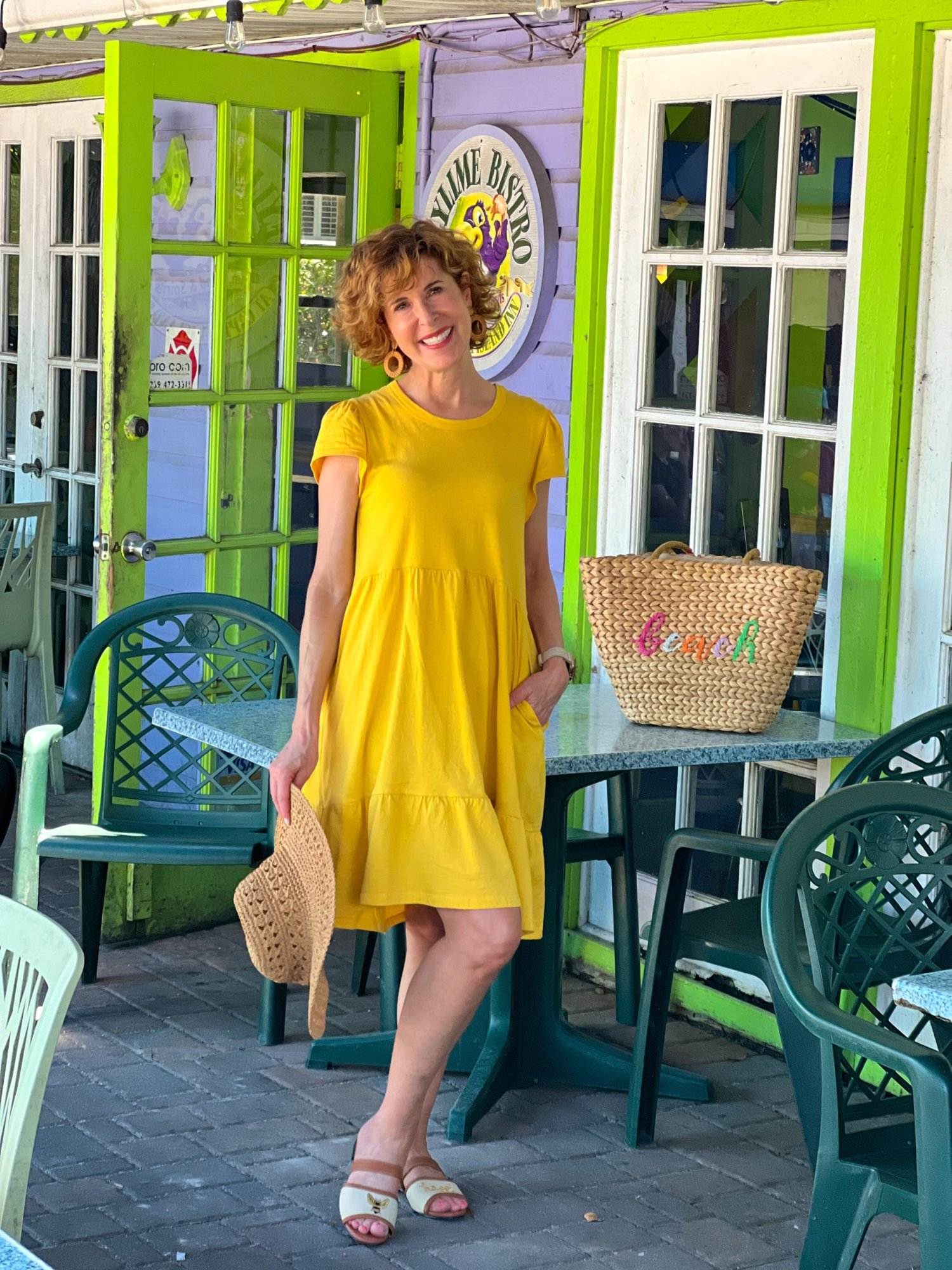 woman over 50 wearing yellow tiered dress holding straw hat with beach bag on table next to her