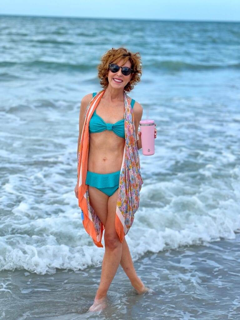 woman over 50 standing pink TAL Stainless Steel Ranger Tumbler on a day in her life at the beach in the surf wearing Time and Tru Women’s and Women's Plus Ruffle High Waist Swim Bottoms with Time & Tru Women's Pareo Swimwear Cover-up on top holding