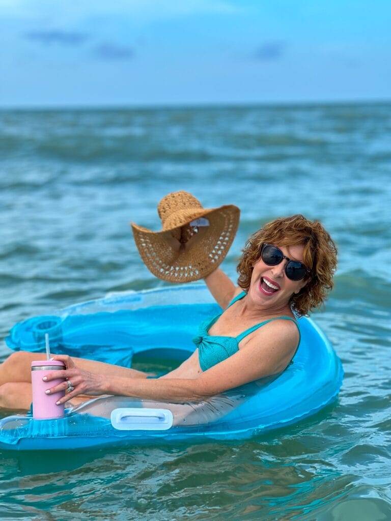 woman wearing sunglasses sitting in Chill-Out Lounge Pool Float from walmart holding a straw hat and pink insulated tumbler with straw on a typical day at the beach