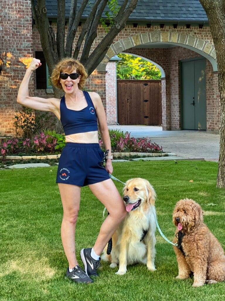 woman standing in front yard with with 2 dogs on leash wearing mighty health sports bra & shorts making a muscle