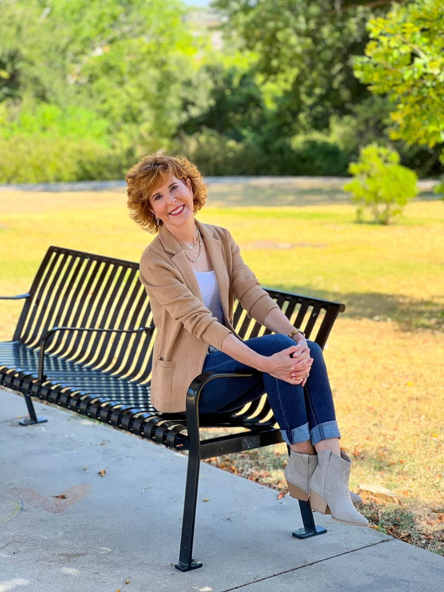 woman wearing western booties, a j.crew factory tan blazer with white cami under it, snake print belt, wit & wisdom ab-solution girlfriend jeans and multiple chains sitting on a park bench with her legs over the side rail