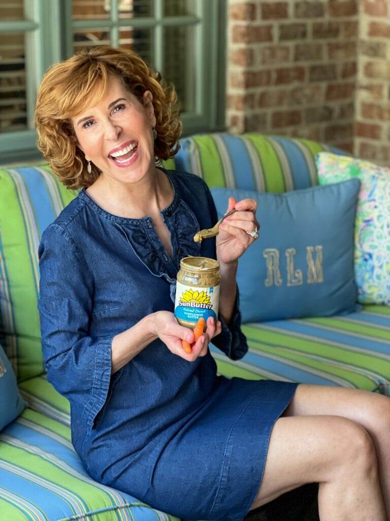 woman wearing blue dress holding sunbutter and carrots sitting on a colorful sofa