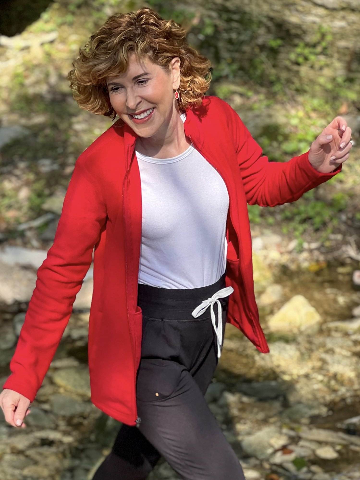 woman wearing lands' end red fleece a white tee and black joggers with red shoes stepping on rocks while crossing a creek bed