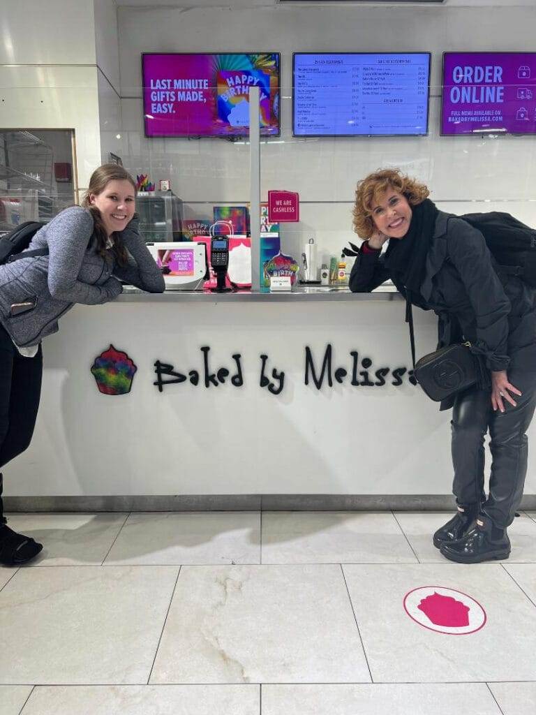 two women standing next to a baked by melissa sign in the store