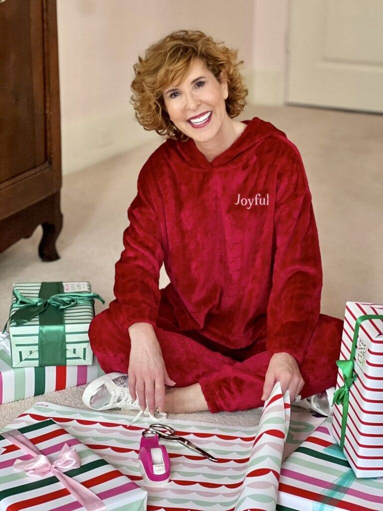 Woman over 50 sitting on floor wearing Joyspun Women's Plush Cable Long Sleeve Hooded 2-piece Top and Pants Pajama Set in red with Joyful written on it