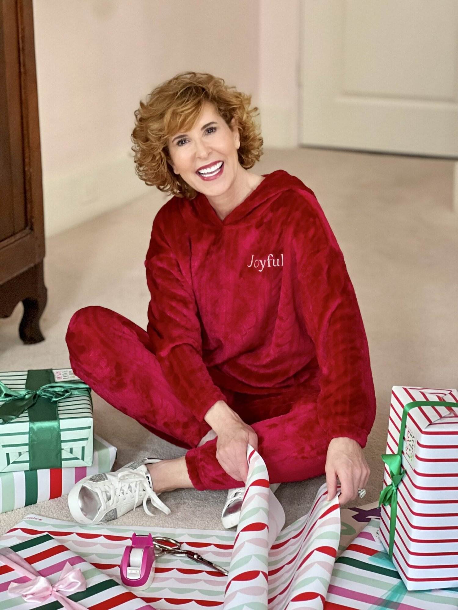 Woman over 50 sitting on floor wearing Joyspun Women's Plush Cable Long Sleeve Hooded 2-piece Top and Pants Pajama Set in red with Joyful written on it