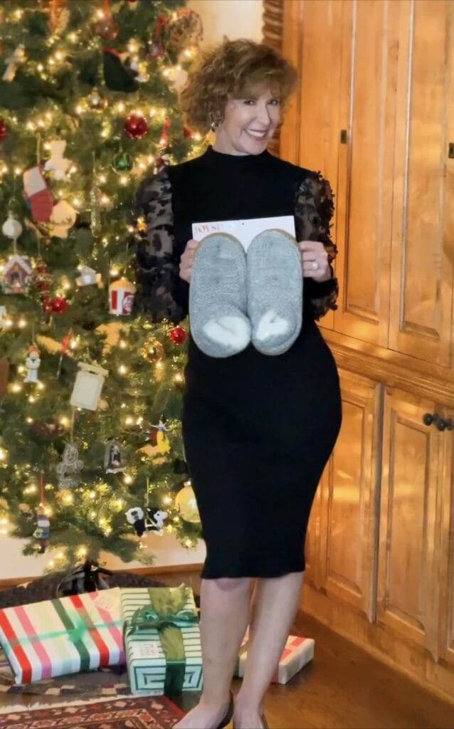 woman over 50 wearing walmart No Boundaries Juniors' Funnel Neck Midi Dress with Sheer Sleeves standing in front of a christmas tree holding a pair of walmart's joyspun slippers