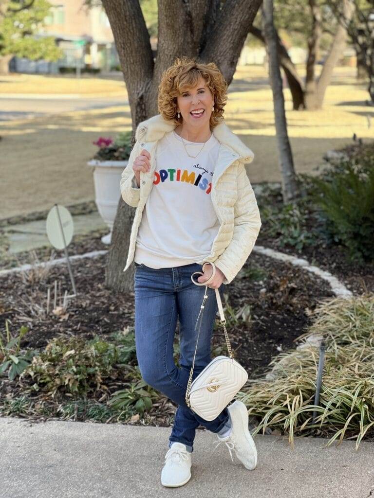 woman over 50 standing on driveway wearing loft's optimist sweatshirt, talbots faux fur collar quilted jacket, jeans, and allbirds sherpa sneakers