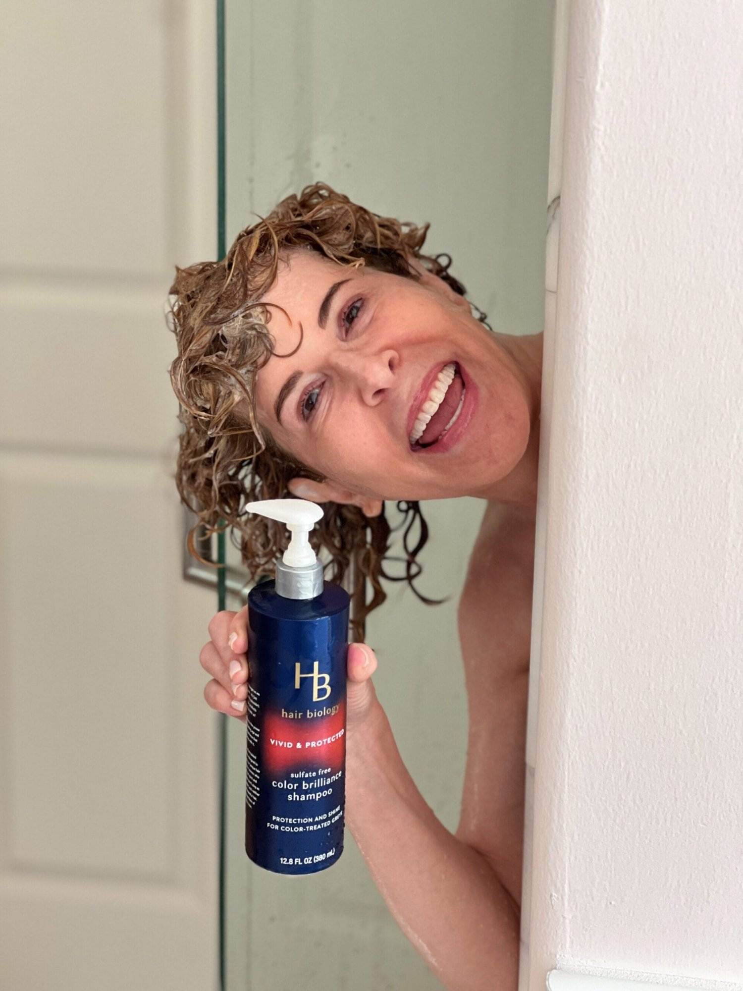 woman peeking out of a shower with shampoo in her hair holding hair biology vivid and protected shampoo