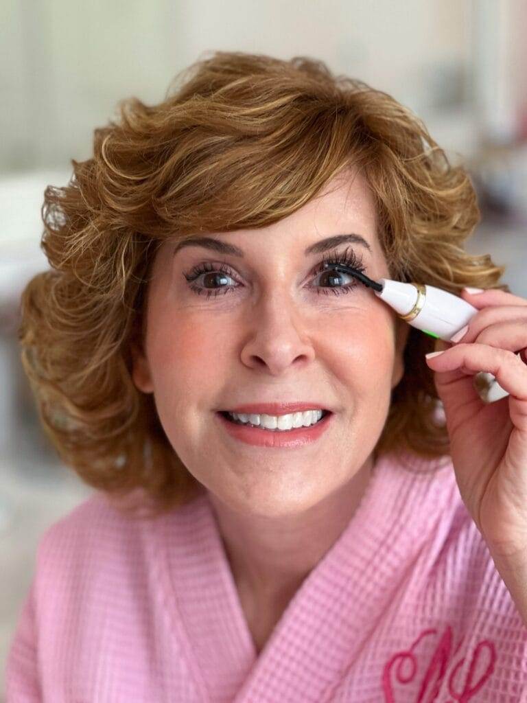 woman over 50 wearing pink robe and looking into camera and using a heated eyelash curler