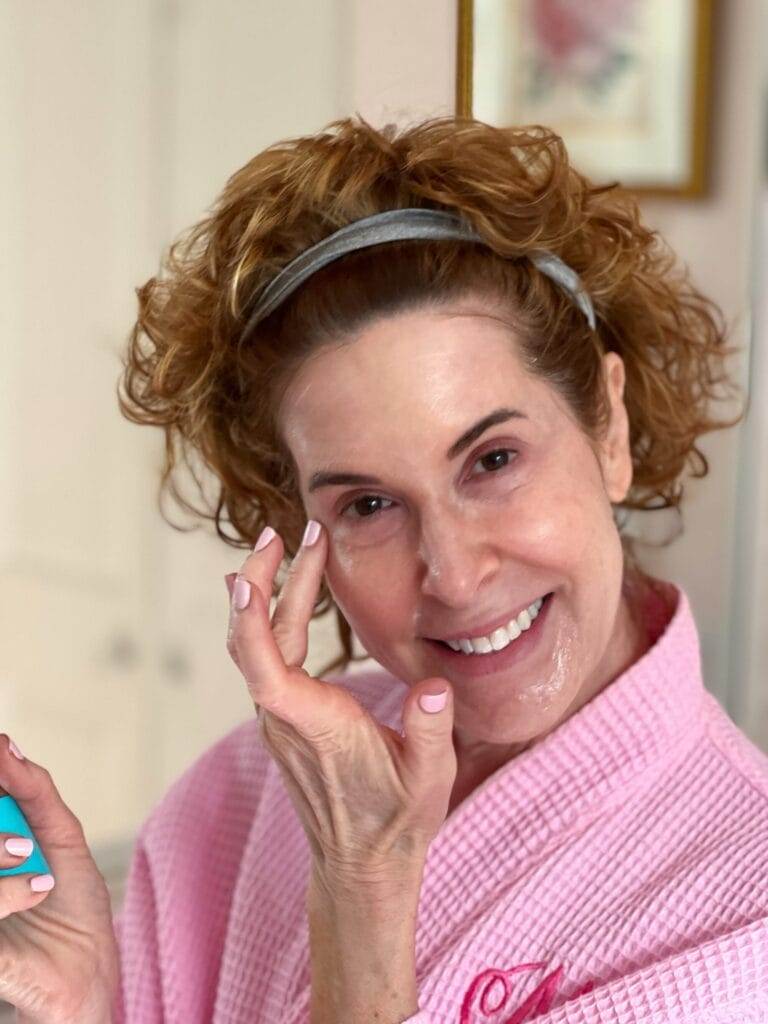 woman over 50 dressed in pink robe applying eye cream as a part of her skincare over 50 regimen