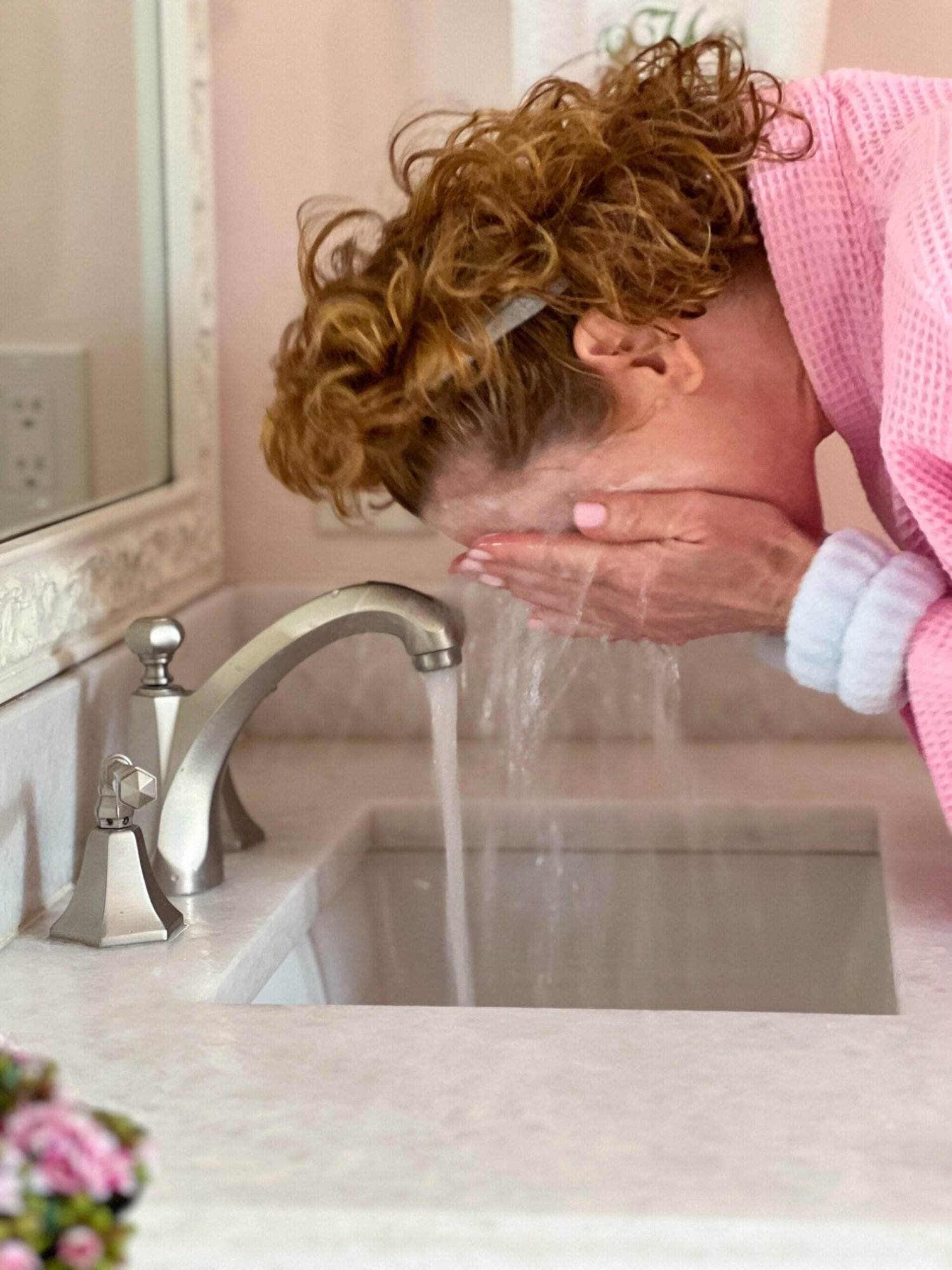 woman over 50 dressed in pink robe rinsing her face