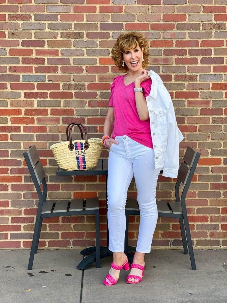 woman holding chico's white peplum denim jacket and wearing white girlfriend jeans with pink amazon v-neck tee and pink target heels standing at a table outdoors next to a handpainted monogrammed straw bag thinking about how to find her purpose in the empty nest