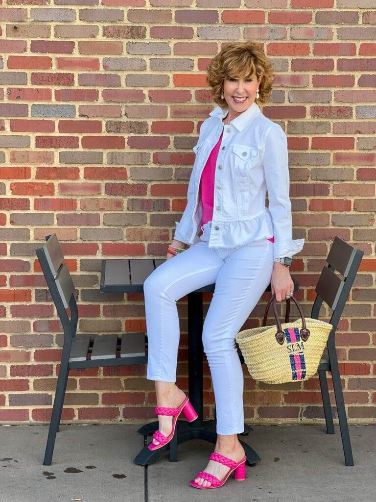 woman wearing chico's white peplum denim jacket and white girlfriend jeans with pink amazon v-neck tee and pink target heels sitting on a table outdoors holding a handpainted monogrammed straw bag