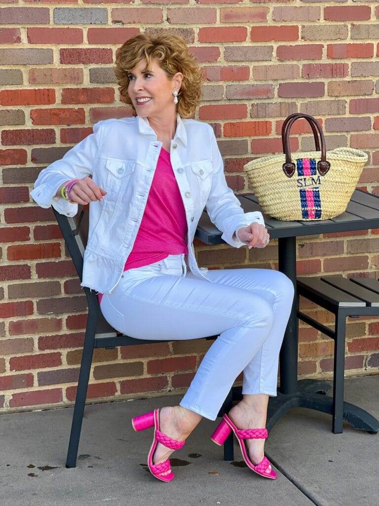 woman wearing chico's white peplum denim jacket and white girlfriend jeans with pink amazon v-neck tee and pink target heels sitting at a table outdoors next to a handpainted monogrammed straw bag