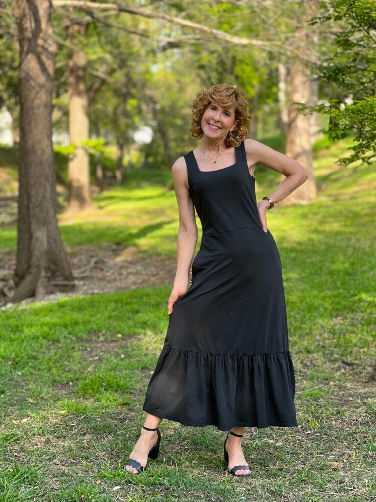 woman with one hand on hip and the other holding out the skirt of her looking into the camera wearing lands' end black tank dress standing in a forest