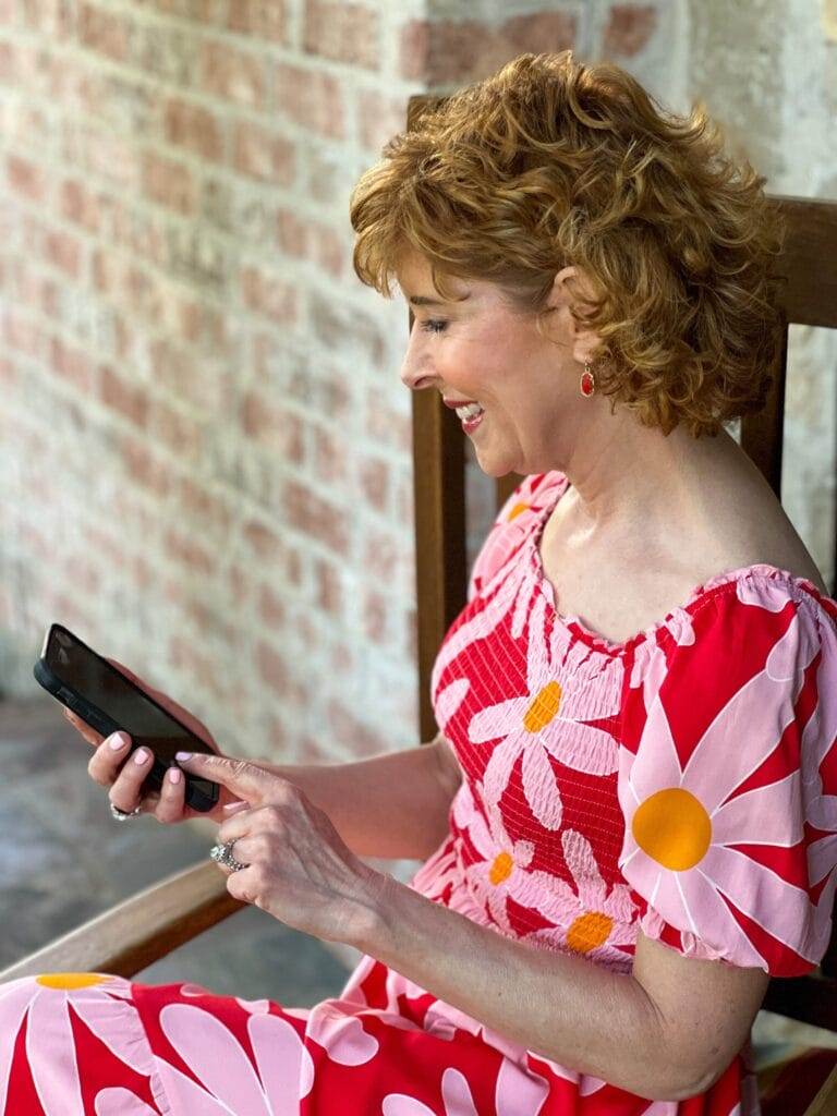 woman wearing red dress with large flowers on it sitting in a chair on her porch looking at her cell phone and smiling