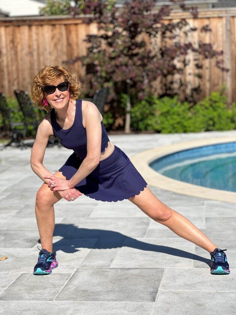 woman over 50 wearing navy blue scalloped workout set standing by a pool demonstrating a side lunge stretch