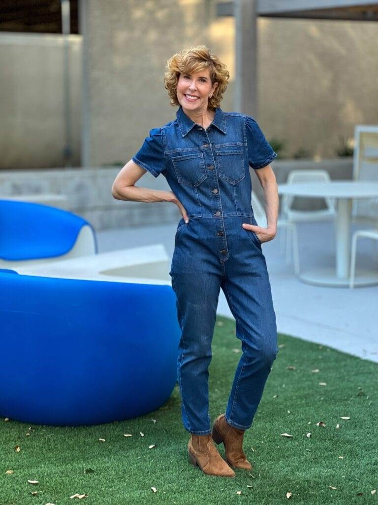 woman over 50 wearing denim puff sleeve jumpsuit with brown western style booties standing on the grass by a blue chair