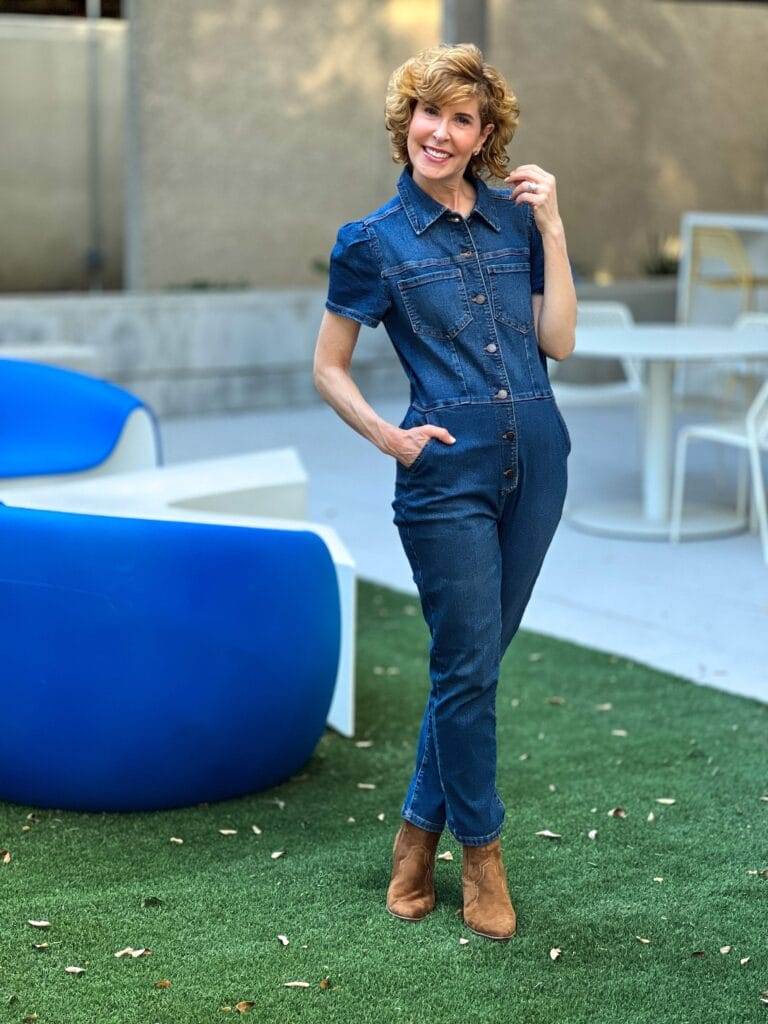 woman over 50 wearing denim puff sleeve jumpsuit with brown western style booties standing on the grass by a blue chair