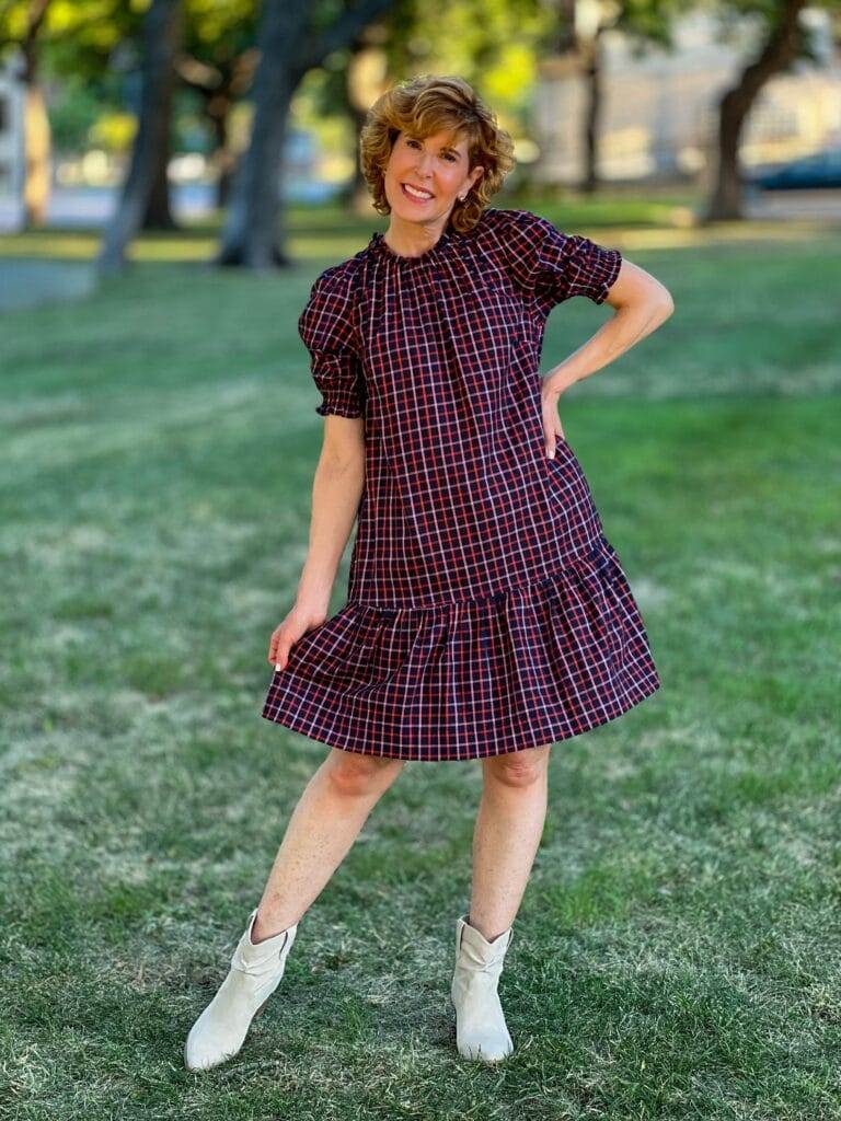 woman over 50 wearing navy plaid shift dress and off white booties standing in the grass