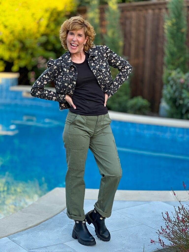 woman standing by a pool wearing green cargo pants, a black tee and a black floral denim jacket with rhinestone studded black booties for fall transition