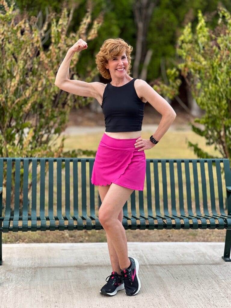 woman over 50 wearing black workout tank and hot pink scallop edge skirt workout wear and making a muscle