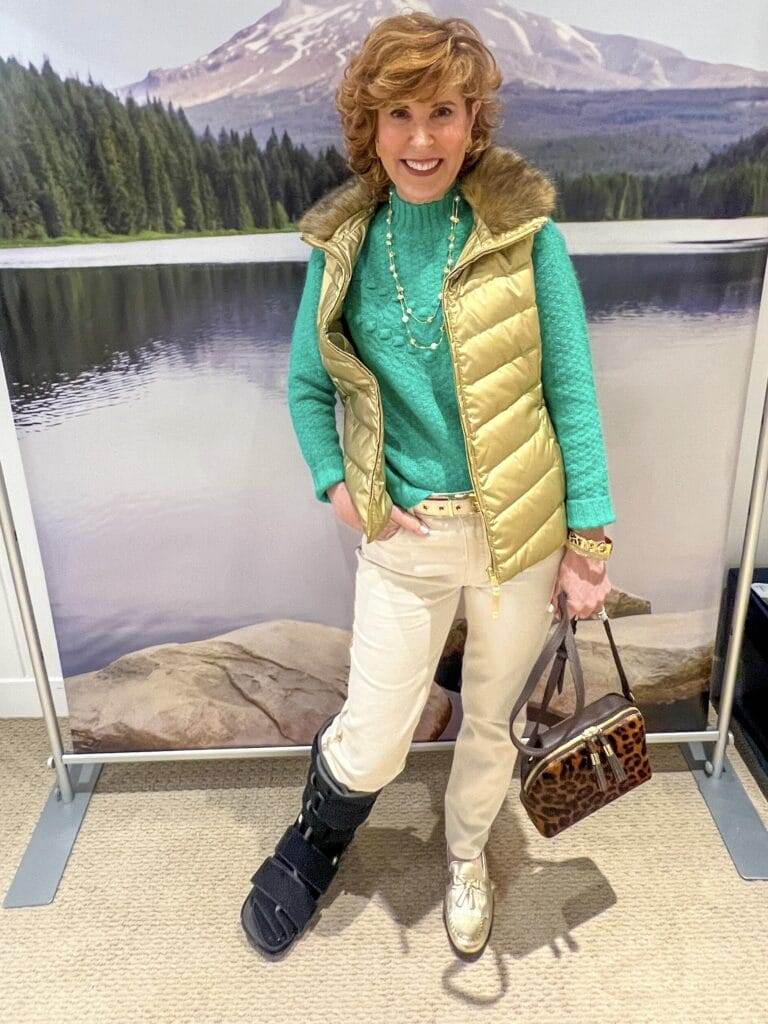 woman posing in front of a snowy backdrop wearing ivory pants, green sweater, and a gold metallic vest