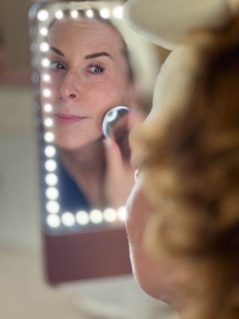 woman over 50 with her hair in a bow headband and wearing a robe using Ziip Halo microcurrent and nanocurrent device while looking into a mirror