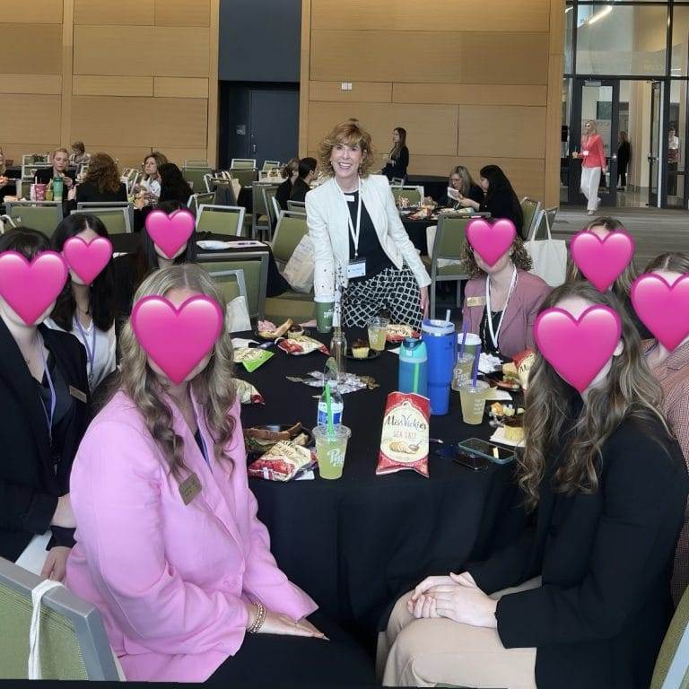 woman standing at the back of a seated group of women with their faces obscured by pink heart for anonymity.