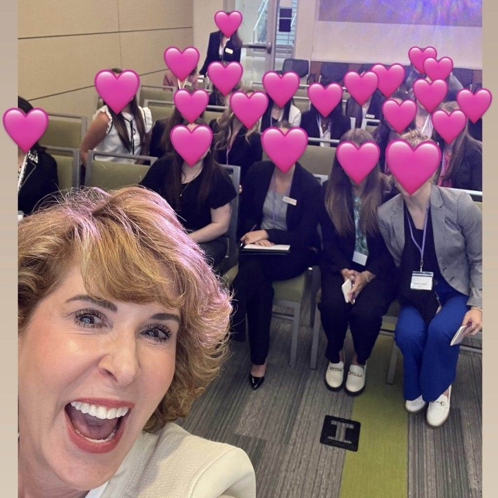 woman standing at the front of a seated group of women with their faces obscured by pink heart for anonymity.