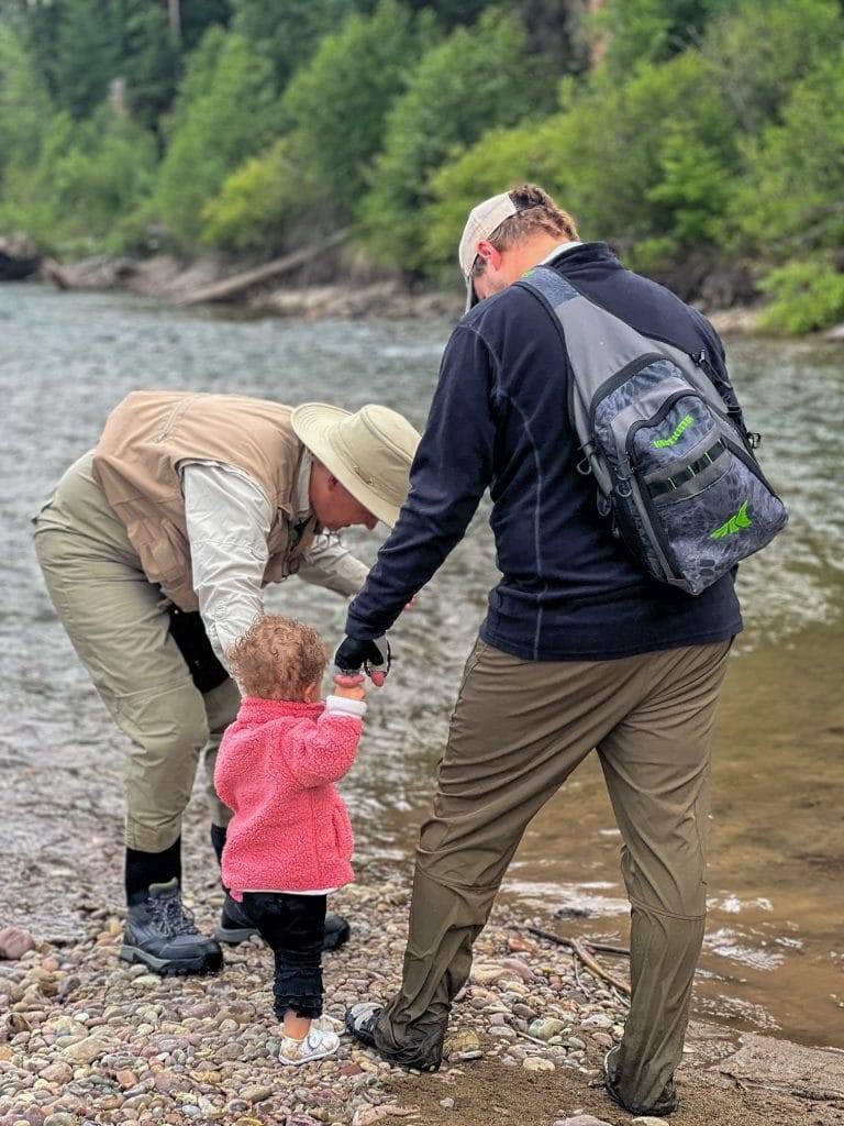 two men in fishing garb and a toddler next to the swan river in montana