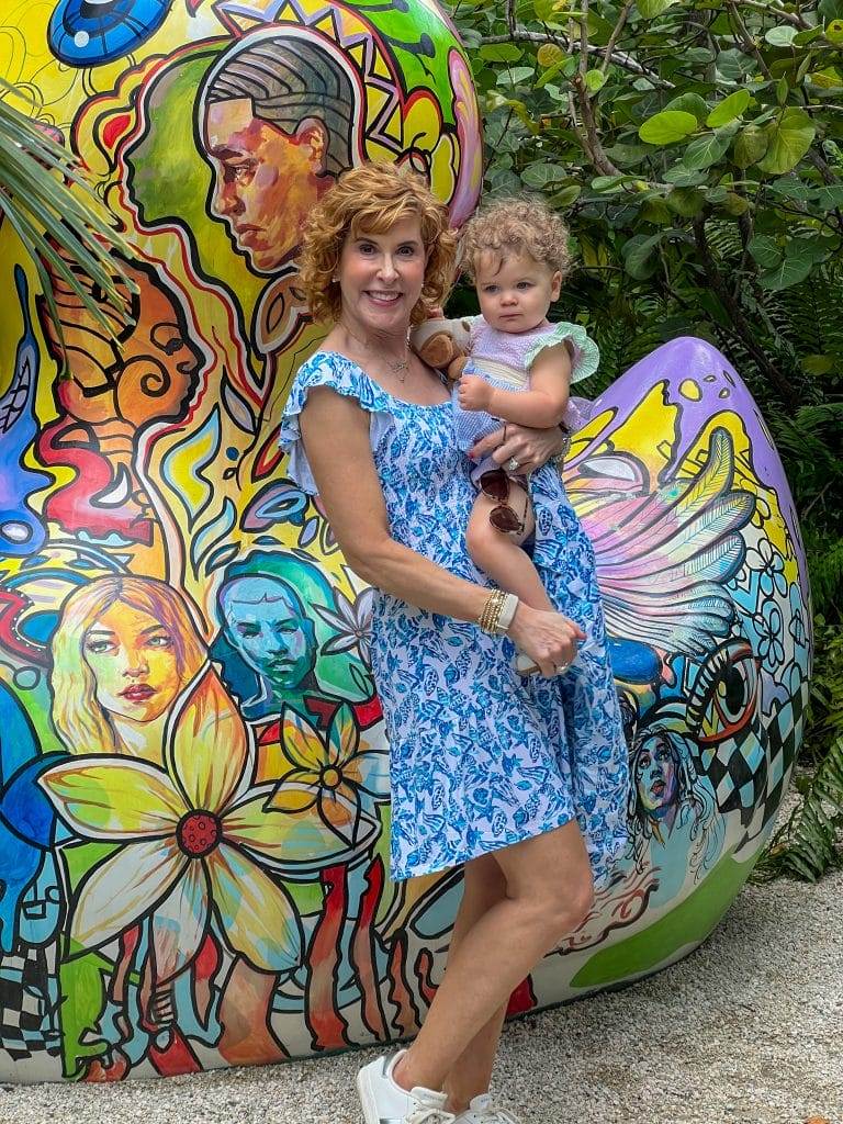 woman wearing lilly pulitzer Jilly Smocked Dress and holding Sea View Linen Button Down Top | Lilly Pulitzer in front of a colorful grafitti rock holding a baby