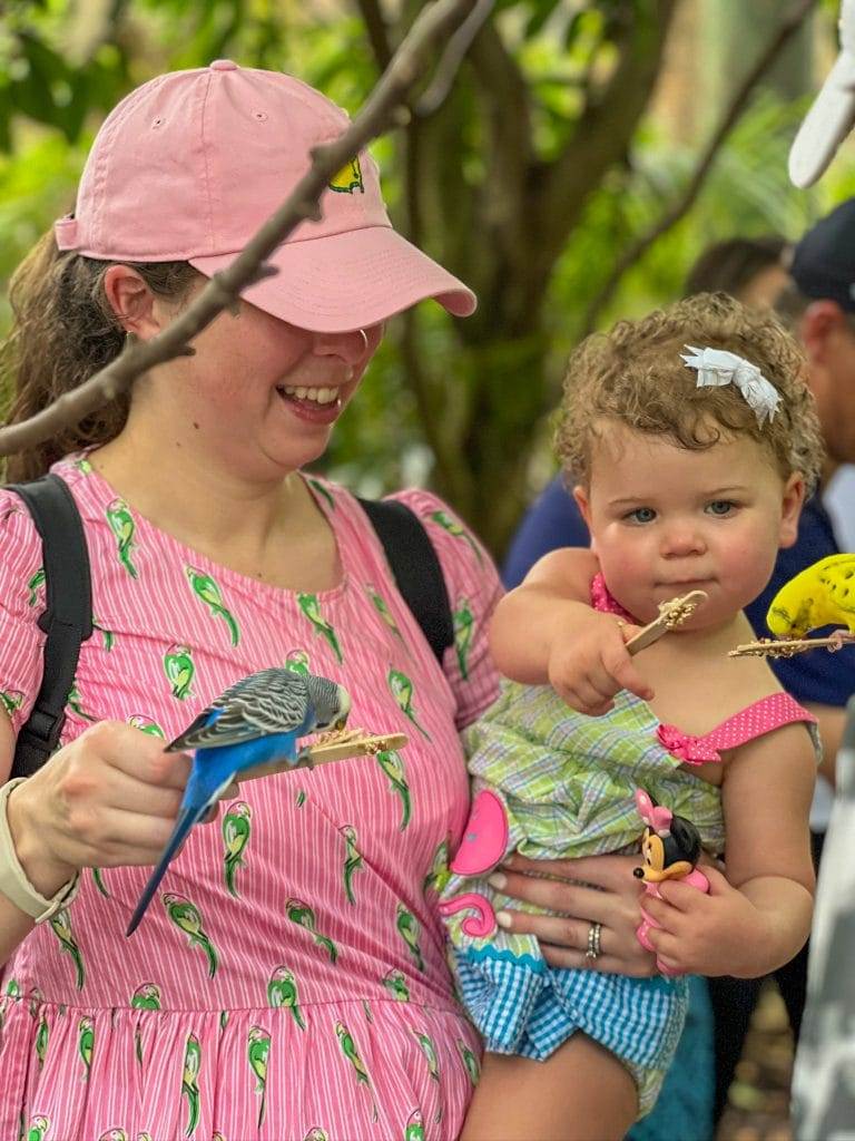 woman dressed in pink holding a baby while they feed colorful birds from birdseed on a stick in Baha Mar