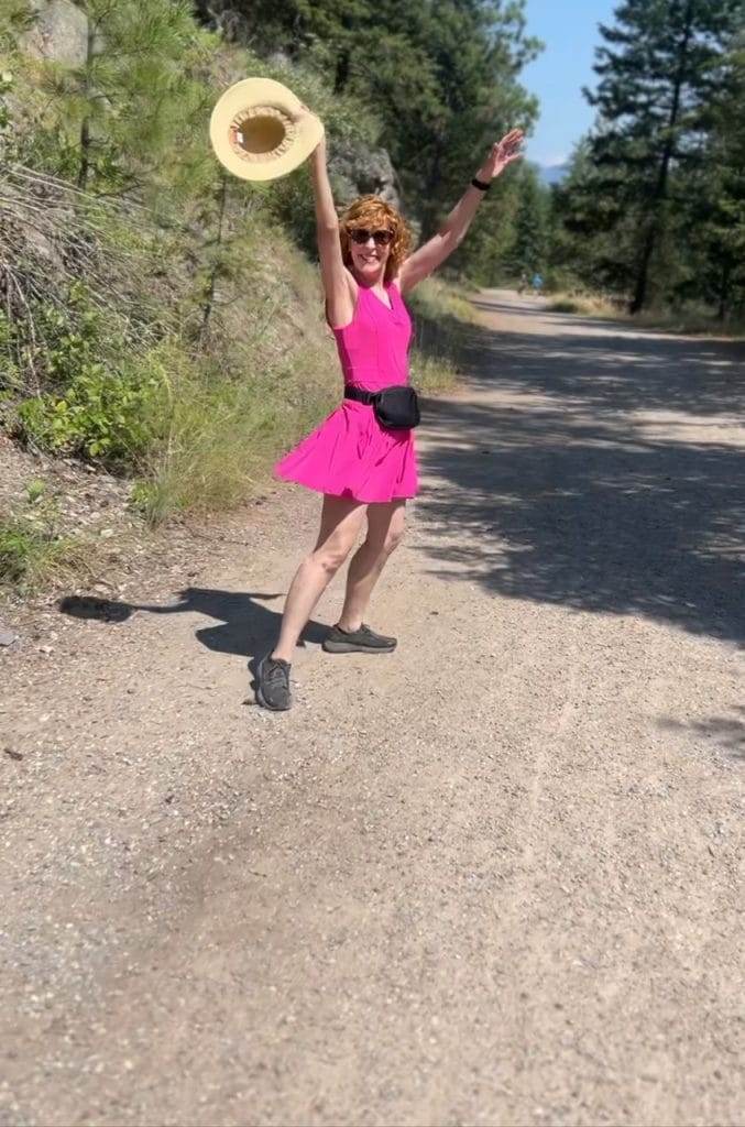 woman wearing pink workout dress walking on a trail holding a hat