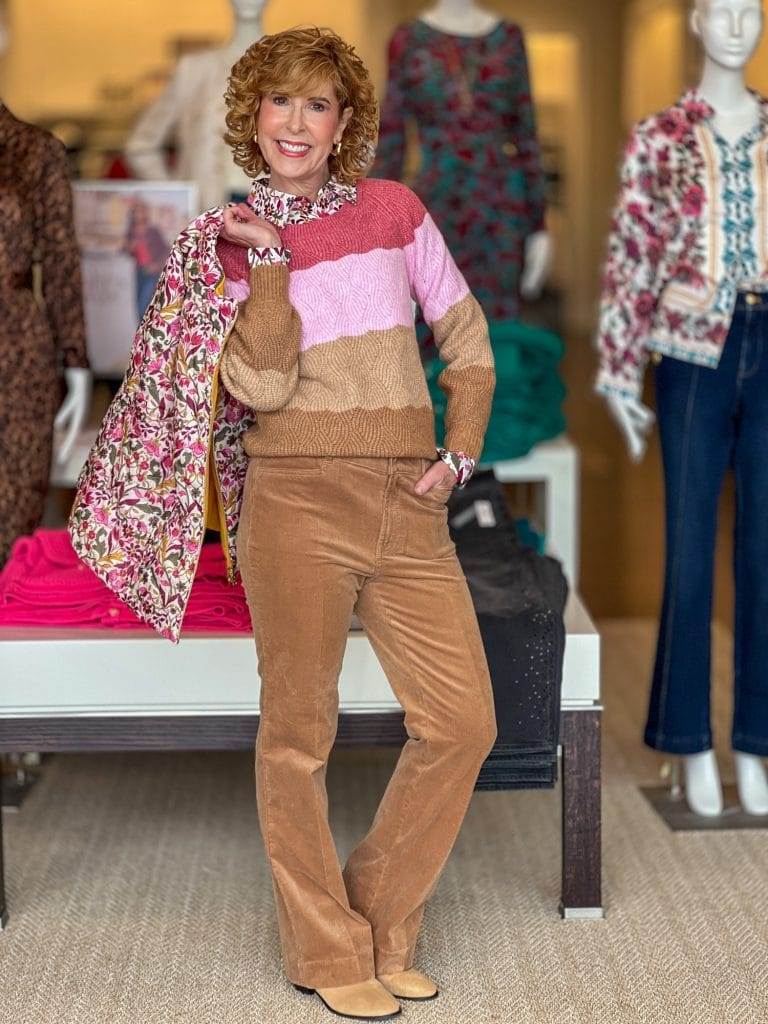 woman over 50 standing in a talbots store wearing brown booties, corduroys, and a striped sweater holding a floral vest over her shoulder