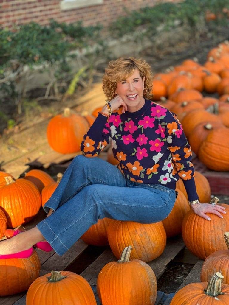 woman sitting on a pumpkin in pumpkin patch with navy blue sweater with pink and orange flowers on it and a pair of wide leg jeans