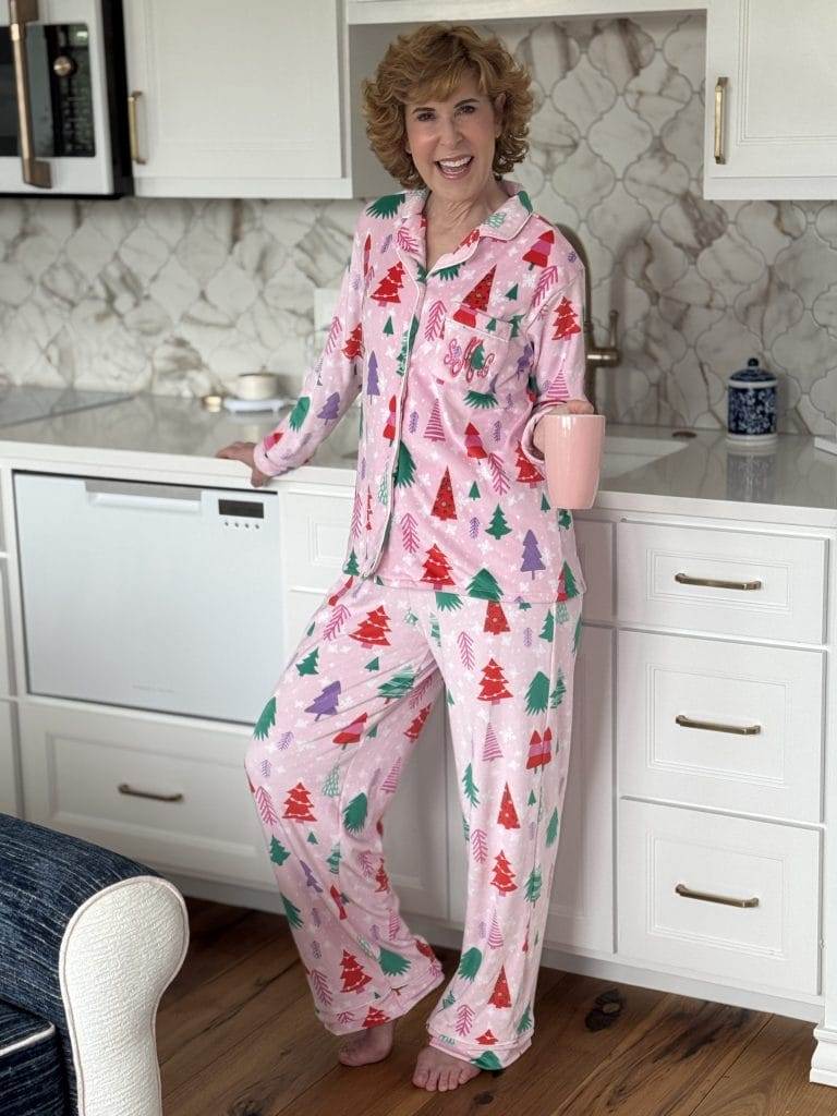 woman standing in a kitchen holding a pink mug in a pink matching pajama set that is long sleeve and long pants with a christmas tree pattern and a monogram on the pocket