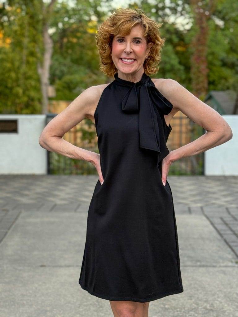 Woman over 50 standing with hands on hips wearing Jude Connally black dress with bow at the neck.