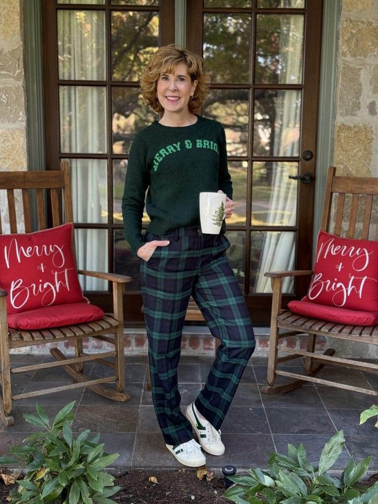 woman over 50 standing on front porch holding spode coffee mug wearing walmart sweater and blackwatch pants with sneakers.