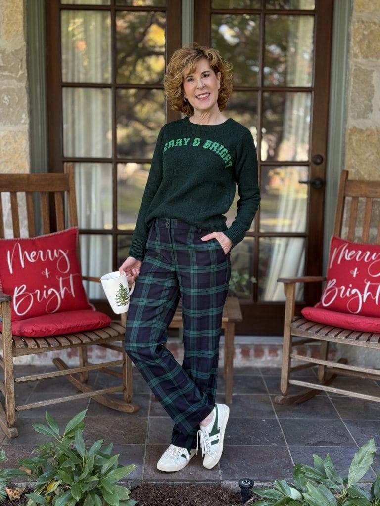 woman over 50 standing on front porch holding spode coffee mug wearing walmart sweater and blackwatch pants with sneakers.