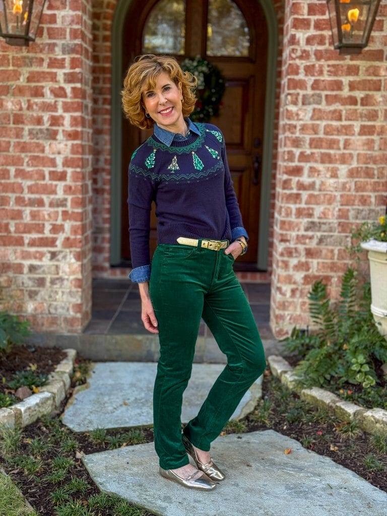 woman standing in front yard wearing green velvet jeans, denim shirt under navy and green christmas tree sweater with gold belt and flats