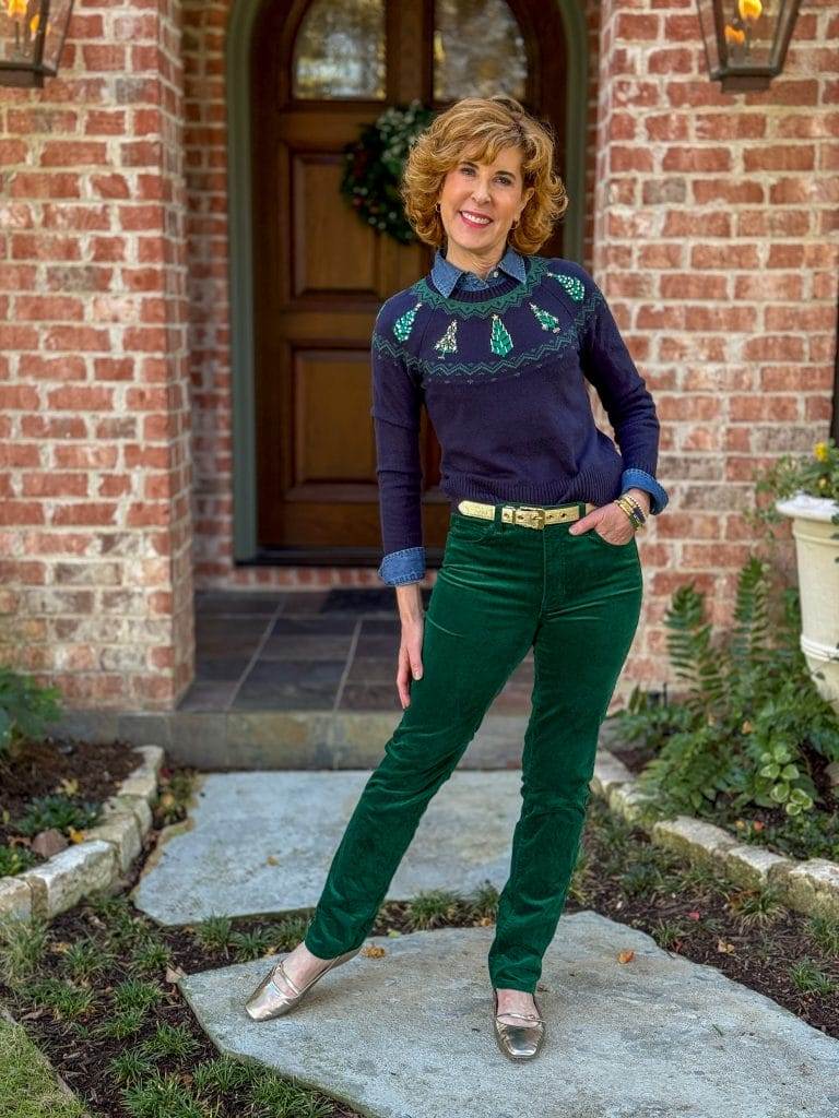 woman standing in front yard wearing green velvet jeans, denim shirt under navy and green christmas tree sweater with gold belt and flats