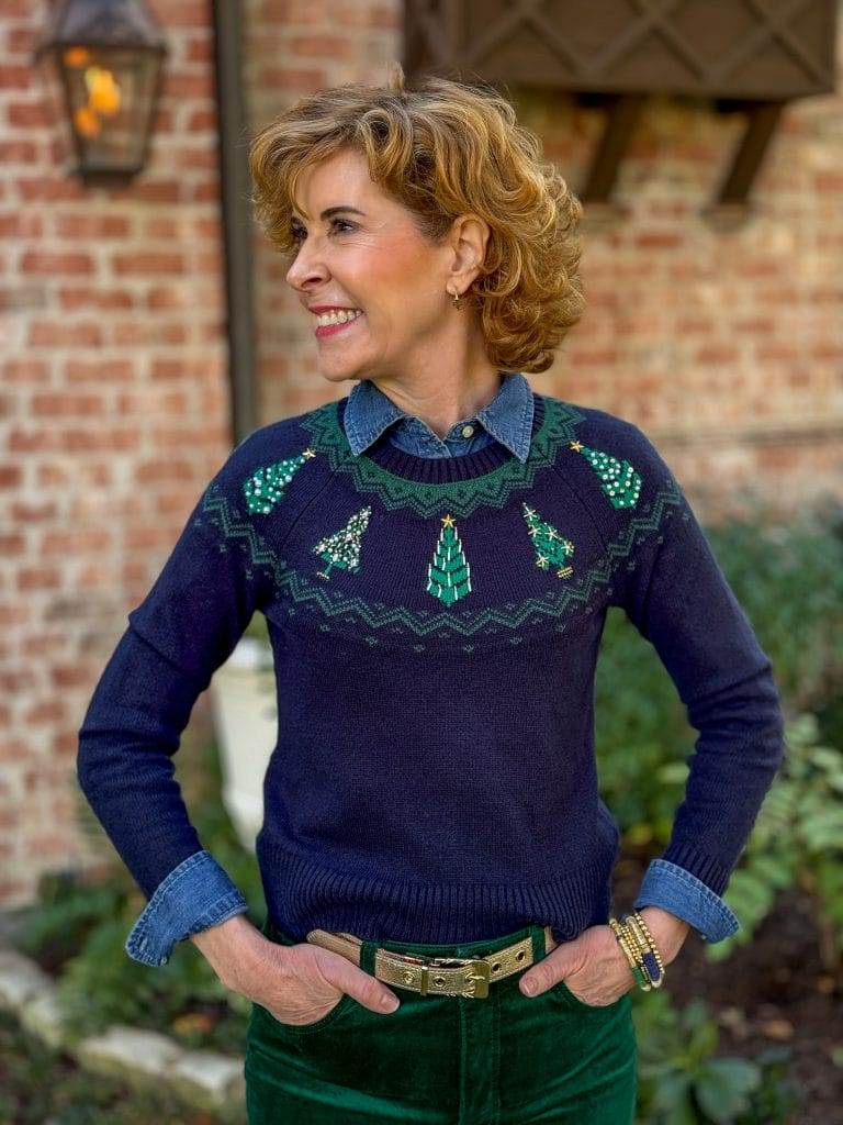 woman standing in front yard wearing denim shirt under navy and green christmas tree sweater with gold belt