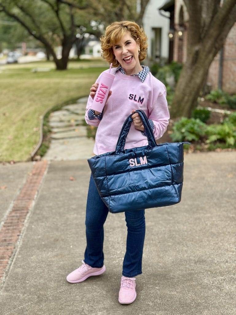 woman wearing sprinkled with pink monogrammed sweat shirt, carrying a pink tumbler with the name SUZY on it, and a navy blue monogrammed puffer bag.