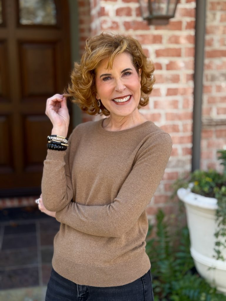 woman over 50 standing in front of a house wearing a mocha mousse cashmere sweater and black and gold bracelets touching her hair